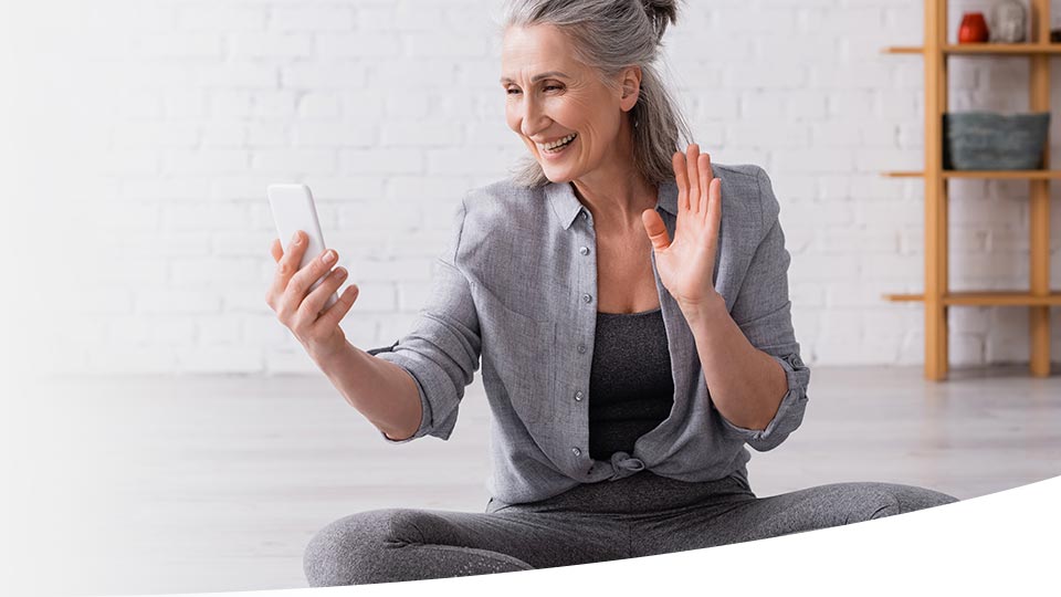 A seated woman holds a smartphone while gesturing with her hand in a modern, minimalistic interior featuring a white brick wall and wooden shelves. No identifiable hearing aids or text present.