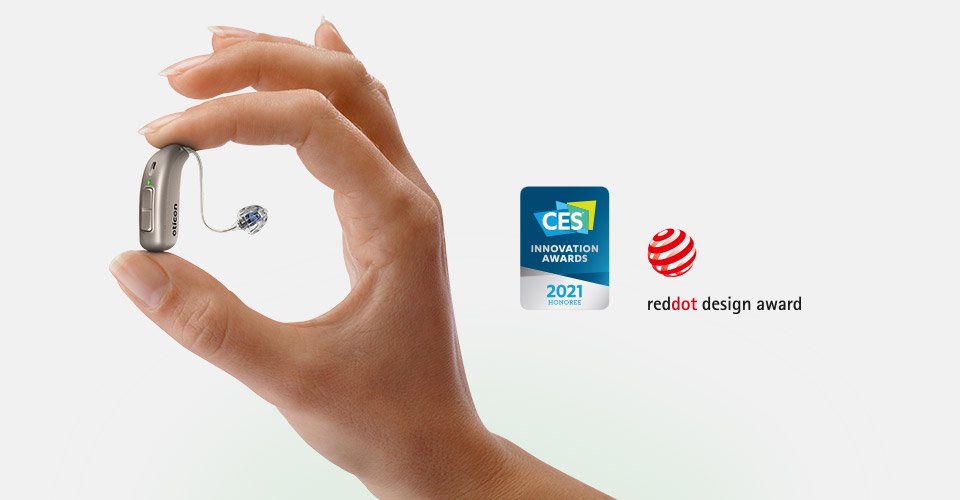 Hand holding a behind-the-ear hearing aid next to CES Innovation Awards and Red Dot Design Award logos