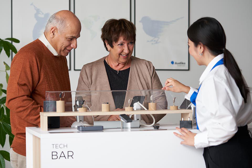 Audiologist demonstrating Bluetooth hearing aids to an older couple at a technology display counter.