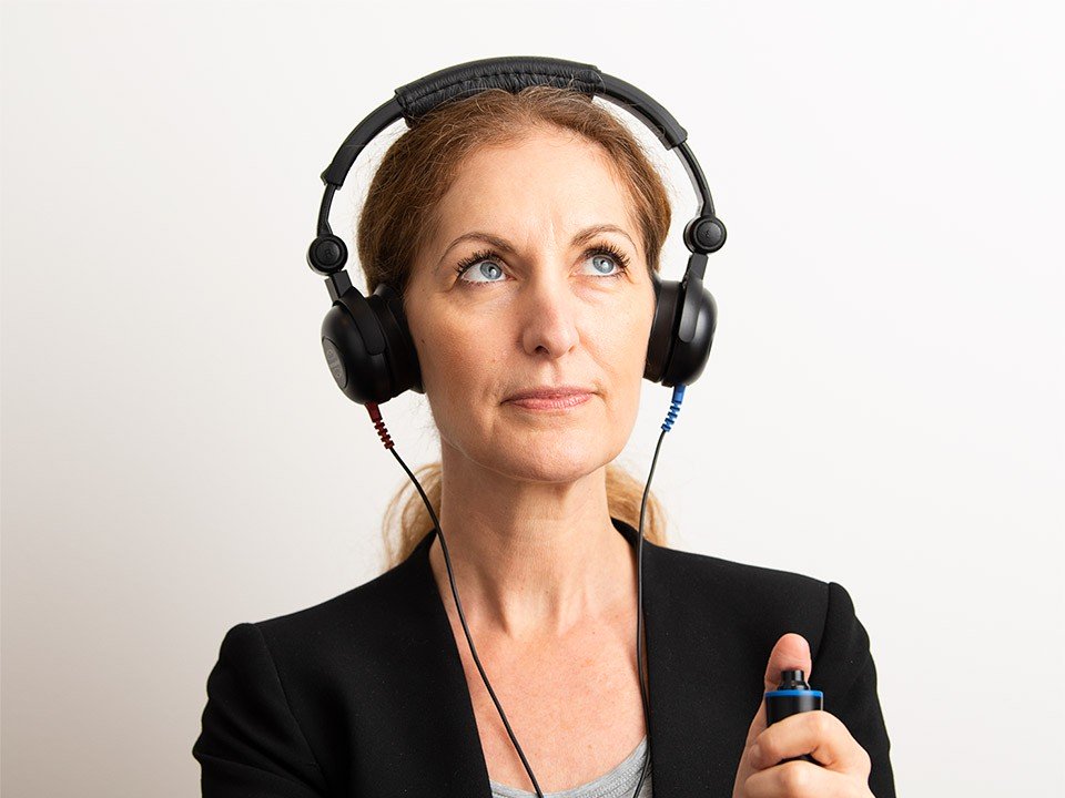 A person wearing over-ear audiometry headphones holds a handheld hearing test response button. The white background and professional attire suggest a clinical setting, possibly a hearing clinic or audiologist session.
