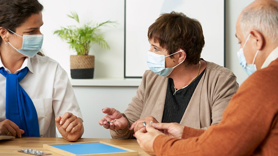 Three people wearing face masks engage in a discussion at a table, with a hearing care professional in a blue scarf assisting a senior couple.