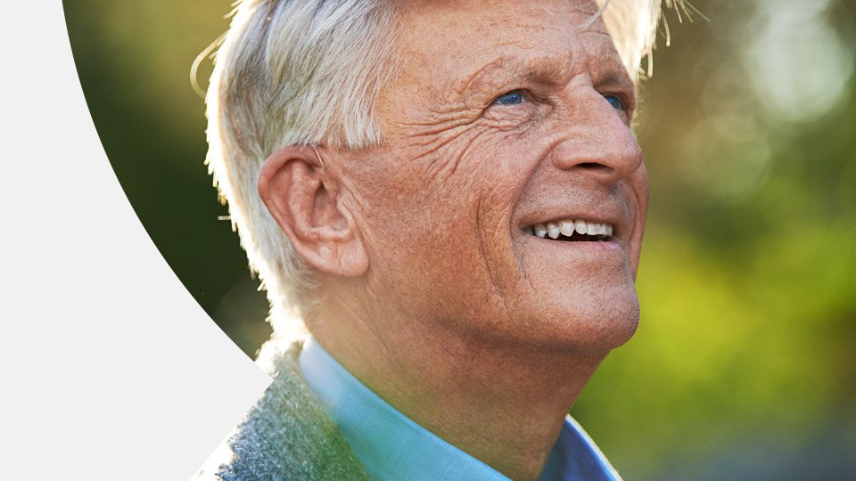 Elderly man with gray hair outdoors, highlighting his ear, with a blurred background in natural light.