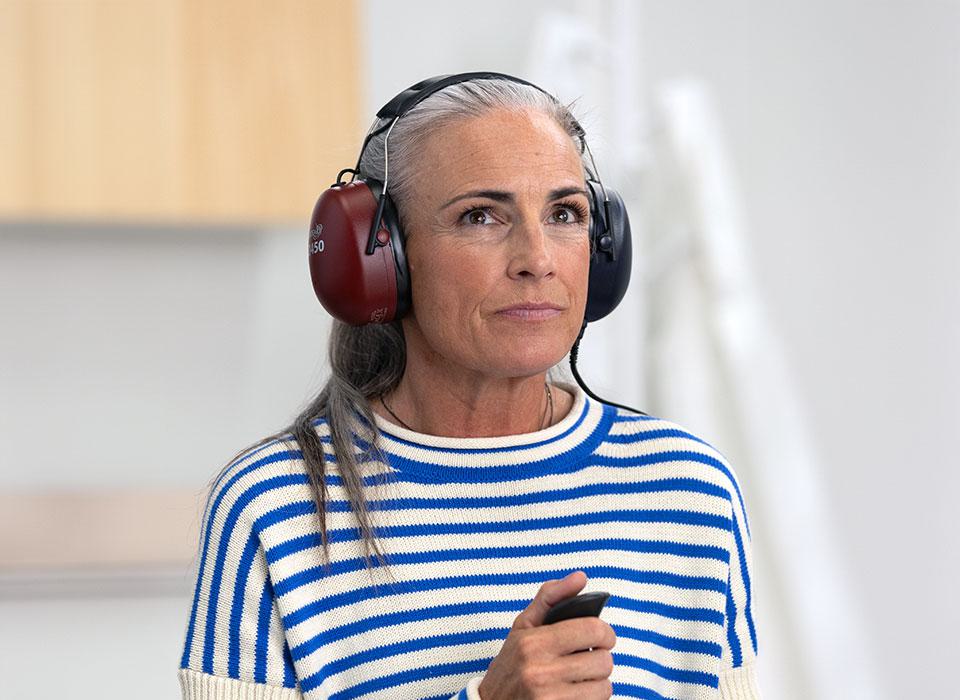 A person wearing noise-canceling earmuffs, holding a handheld device, stands indoors in a brightly lit room with white walls and blurred furniture, suggesting a hearing test environment. Text on earmuffs reads ’Peltor Optime III.’