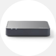 A black and gray rectangular Oticon device lays flat against a plain white background, designed for hearing aid connectivity and streaming, with 'Oticon' branded subtly on the top surface.
