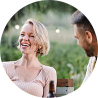 A woman with short blonde hair, wearing a light pink top, engages in conversation with a man outdoors in a garden setting, featuring blurred string lights and greenery in the background.