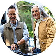 Two individuals, a man and a woman, stand outdoors, with blurred faces suggesting anonymity. The man wears a brown vest over a gray shirt, and the woman wears a mustard quilted jacket over a cream turtleneck. They are positioned near a bicycle, surrounded by a lush, green setting, indicating a casual outdoor activity like cycling.
