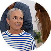 An older adult wearing a blue and white striped shirt sits in a hearing clinic, engaged in discussion with an audiologist. A blurred face obscures the individual's identity in a professional setting.