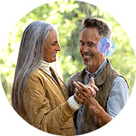 A couple dances outdoors in warm sunlight, holding hands affectionately. The man wears a discreet, behind-the-ear hearing aid, likely highlighting hearing solutions available at Audika hearing clinics.