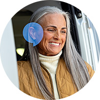 A senior individual with long gray hair, wearing a brown quilted jacket and cream turtleneck, stands by a vehicle door. A blue overlay highlights the ear region, implying hearing aid usage.