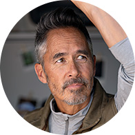 A middle-aged individual adjusts their hair in a casual indoor setting with blurred background elements, including photo frames. No visible branding or hearing aids are identifiable in the image.