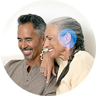 A woman wearing a discreet blue hearing aid rests her arm on a man's shoulder, seated on a couch in a well-lit setting, symbolizing care and hearing health solutions.