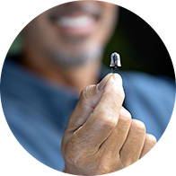 A smiling man holds a small, discreet hearing aid between his fingers, showcasing its compact design against a blurred background, suggesting focus on advanced hearing care technology and user-friendly devices.