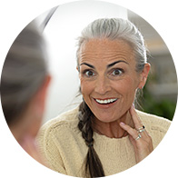 A woman with gray hair in a braid wears a cream sweater, sitting indoors during a conversation. A blurred second figure faces her, suggesting a consultation, possibly at a hearing clinic. No text is visible.