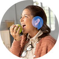 A woman with long brown hair wears a discreet hearing aid while holding an apple near her mouth. She is indoors, dressed in a knit cardigan and glasses hang from her collar.