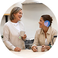 Two women are conversing in a kitchen, each holding a mug; one wears a discreet blue hearing aid on her ear, promoting hearing care solutions. A modern and casual environment surrounds them.
