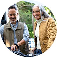 Two individuals, a man and a woman, stand outdoors, with blurred faces suggesting anonymity. The man wears a brown vest over a gray shirt, and the woman wears a mustard quilted jacket over a cream turtleneck. They are positioned near a bicycle, surrounded by a lush, green setting, indicating a casual outdoor activity like cycling.