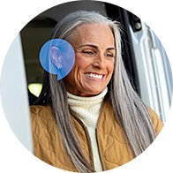 A senior individual with long gray hair, wearing a brown quilted jacket and cream turtleneck, stands by a vehicle door. A blue overlay highlights the ear region, implying hearing aid usage.