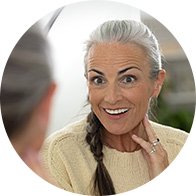 A woman with gray hair in a braid wears a cream sweater, sitting indoors during a conversation. A blurred second figure faces her, suggesting a consultation, possibly at a hearing clinic. No text is visible.