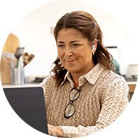 A woman wearing a hearing aid sits in a kitchen environment, working on a laptop. Reading glasses rest on her sweater, and kitchen utensils and appliances are visible in the background.