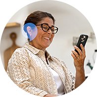 A woman wearing glasses and a beige blouse uses a smartphone while seated in a home setting. A blue hearing aid, possibly from Oticon, is visible behind her ear.