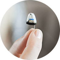A hand holds a tiny Oticon hearing aid with a discreet, clear design, set against a softly blurred background, emphasizing innovative hearing care technology and precision from Audika's hearing clinic.