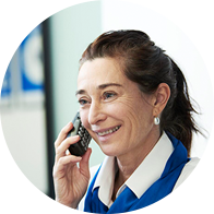 A woman wearing a white collared shirt and blue scarf is on a phone call in a professional setting, possibly a hearing clinic. A blurred background shows indistinct signage.