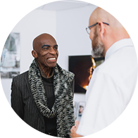 A bald audiologist wearing glasses converses with a patient dressed in a patterned scarf in a modern hearing clinic. Wall-mounted visuals and white decor complete the professional setting. No visible text.