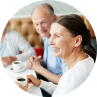 A group of middle-aged individuals, seated at a table in a casual café setting, hold cups of coffee while conversing. The setting suggests a relaxed environment fostering social interactions. No visible text or hearing aid brands are identifiable.