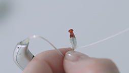 A silver behind-the-ear hearing aid with a clear tube is held by a hand, focusing on the red-tipped earbud. The background is softly blurred, emphasizing the device details.