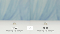 Two small, cylindrical hearing aid batteries sit on a white surface against a blurred blue background. The left battery is labeled 'NEW Hearing aid battery,' and the right is labeled 'OLD Hearing aid battery,' separated by 'vs.'