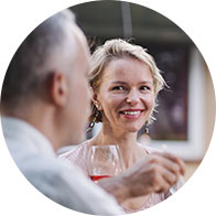 A gray-haired man holds a wine glass and converses with a blonde woman (blurred face) in a warmly lit setting, suggesting a social or dining experience. No text visible.
