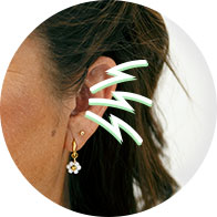 A close-up of a woman's ear wearing a gold and daisy-shaped earring, illustrated with green sound waves to symbolize hearing or sound perception, set against a neutral background. No text present.