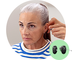 A hearing care professional fits a discreet hearing aid behind an older woman's ear in a clinical setting. Nearby, a graphic displays black Oticon hearing aids with green accents.