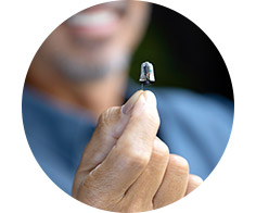 A hand holds a discreet in-the-ear hearing aid, emphasizing its small size. The background shows a smiling individual, slightly blurred, suggesting satisfaction with the device in a hearing care context.