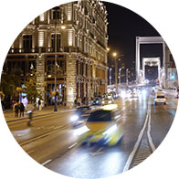Busy city street with moving cars and blurred headlights at night, bordered by an illuminated historic building on the left and a modern bridge in the background. No text is visible.
