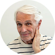An older adult with white hair, wearing a striped shirt, touches their ear, possibly referencing hearing issues. The plain background suggests a setting like a hearing clinic or consultation. No visible hearing aids, text, or identifiable branding.