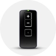 A sleek black remote control with three buttons (+, -, and central square), featuring a green indicator light at the top, designed for adjusting hearing aids in a hearing care setting.
