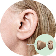 A close-up of a woman’s ear wearing a discreet in-the-ear hearing aid. Next to the ear, a detailed inset shows two styles of hearing aids in beige.