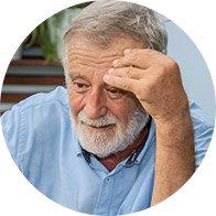A senior individual wearing a light blue shirt rests their head on one hand. The background features blurred greenery and elements suggesting an outdoor or patio setting. No identifiable hearing aids, text, or brand markings are visible.