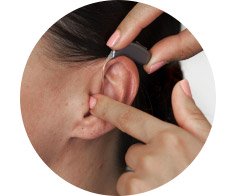 A behind-the-ear hearing aid is being fitted on a woman’s ear by hands with neatly manicured nails, emphasizing hearing care services in a clinical or personal setting. No text visible.