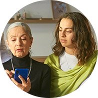 Two individuals, with blurred faces, interact; one holds a blue smartphone. They sit in a well-lit, cozy environment with visible wooden shelves and decorative items in the background. No text is present.