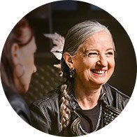 Two individuals are seated indoors, engaged in conversation. One has long gray hair in a braid and wears a leather jacket; the other is partially visible. The environment appears dimly lit and casual. No text or identifiable hearing aid brands are visible.