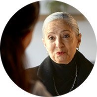 A person with short, silver hair and blurred facial features wears a black shirt and jewelry, sitting indoors across from another individual, likely in a consultation or hearing clinic setting.