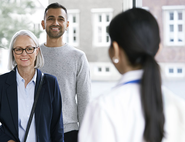 A white-haired woman and a younger man stand facing a professional audiologist in a white coat inside a hearing clinic. The clinic's bright interior has large windows showing urban buildings.
