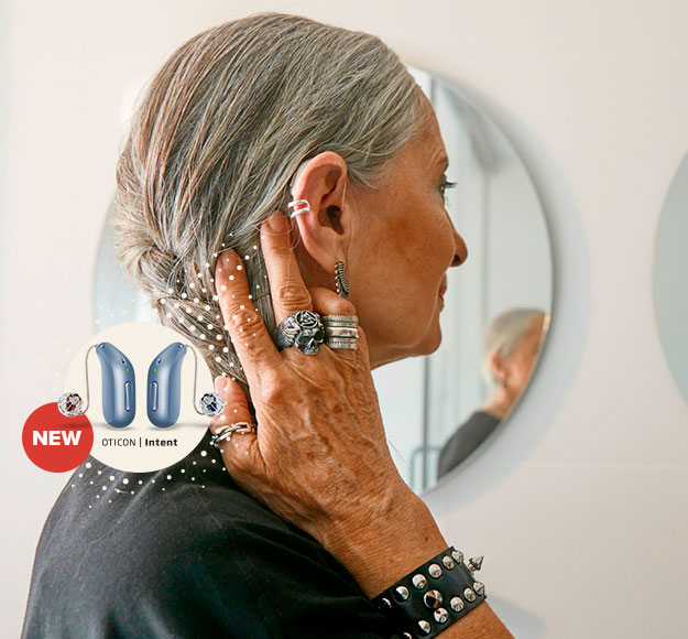 A person with silver hair adjusts a hearing aid behind their ear while standing in front of a circular mirror. The image highlights ’Oticon | Intent’ hearing aids with a ’NEW’ label. The surroundings suggest a modern bathroom or personal care setting.