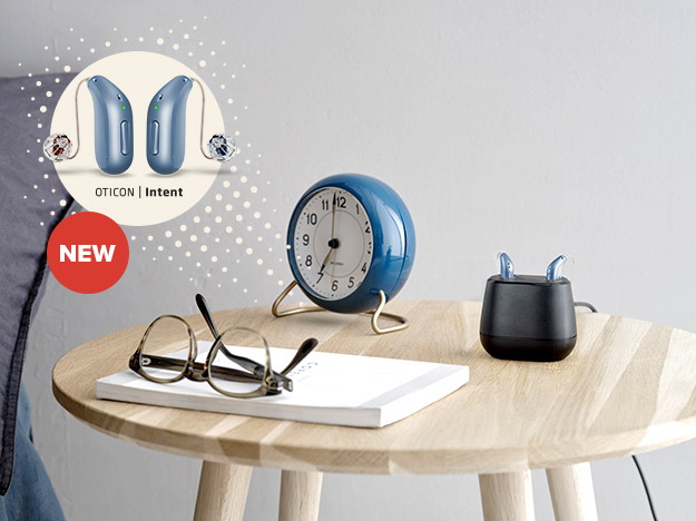 A pair of Oticon Intent hearing aids rest in a black charging station on a wooden table beside a blue alarm clock, glasses, and a notebook. Text reads: 'NEW OTICON | Intent.'