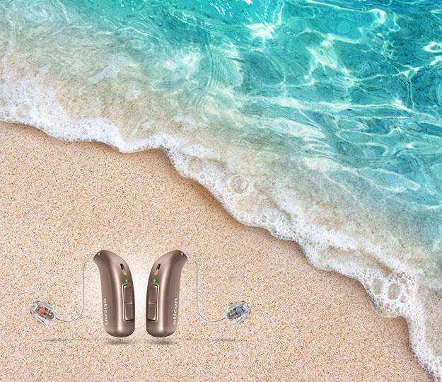 Two Oticon hearing aids resting on sandy beach sand as gentle ocean waves approach, emphasizing advanced hearing technology in a serene, natural outdoor setting. Text on the devices reads ’Oticon.’