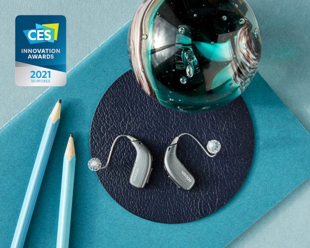 Behind-the-ear hearing aids placed on a textured surface alongside blue pencils, a decorative glass orb, and a CES Innovation Awards 2021 Honoree badge.