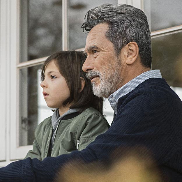 An older man, with short gray hair, sits closely beside a child wearing a green jacket, near a large window in a cozy domestic environment. No visible hearing aids or text are present.