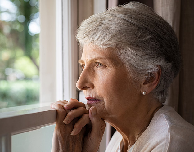 Older woman with short grey hair looking out of a window, hands resting on the window frame, sunlight illuminating the background.